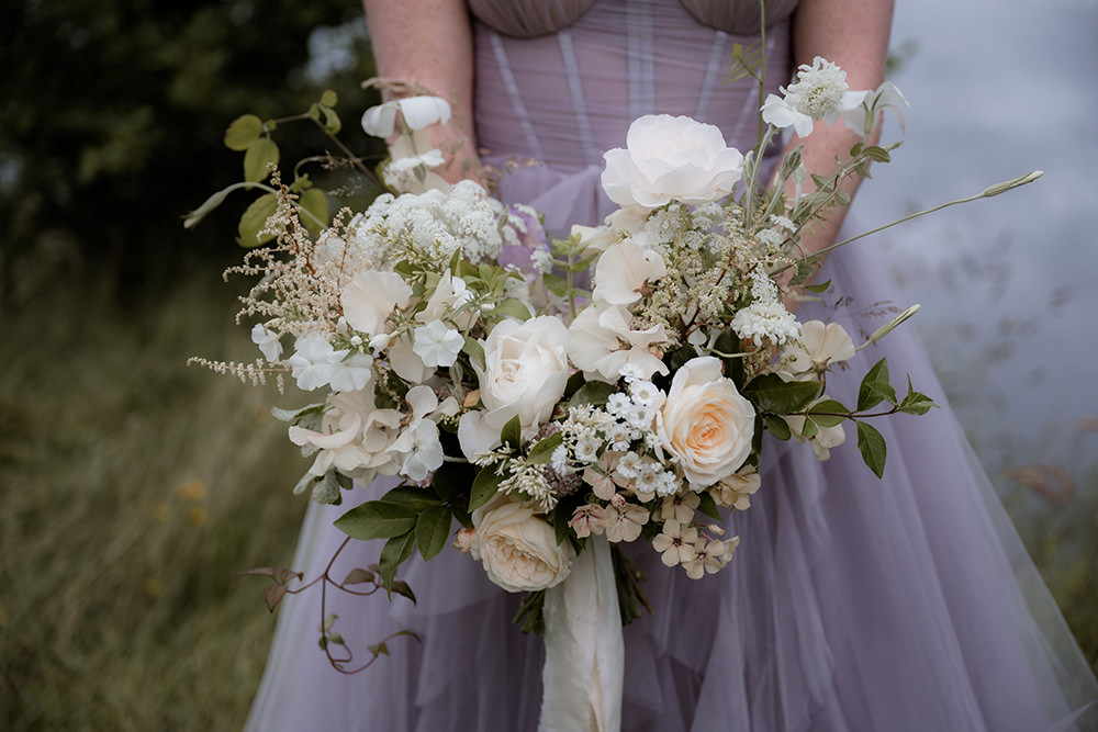 Close-up of a natural, garden-style bridal bouquet with white and cream roses, sweet peas, and wild greenery, held against a soft lavender tulle wedding dress