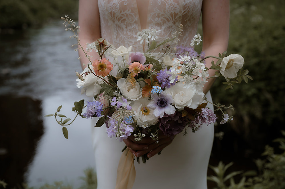 Colorful wildflower-inspired bridal bouquet featuring white roses, anemones, scabiosa, and delicate pastel blooms, held in front of a lace wedding dress near a river