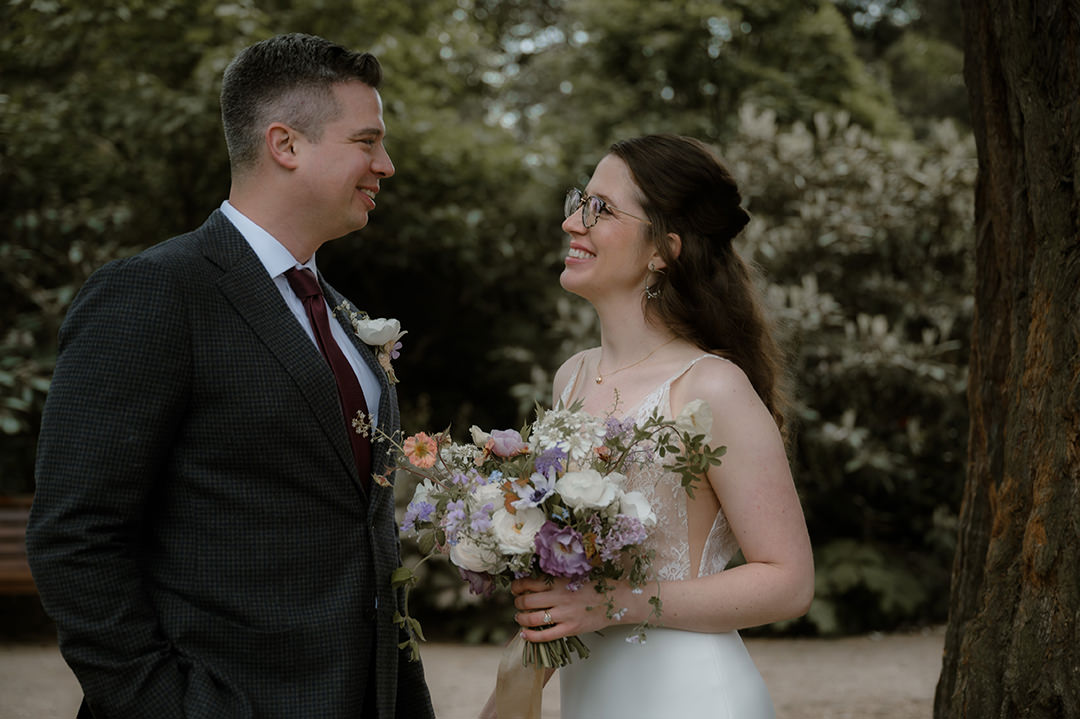 Bride in a lace-backed gown smiles at her groom while holding a pastel bouquet of roses and wildflowers against a backdrop of lush greenery at the Edinburgh Botanics