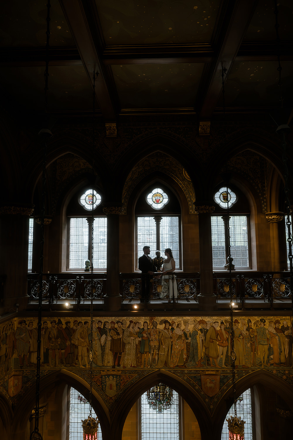 A couple stands on an ornate balcony inside a grand historic building, framed by large arched windows with stained glass and intricate architectural details, above a mural of figures in medieval-style attire