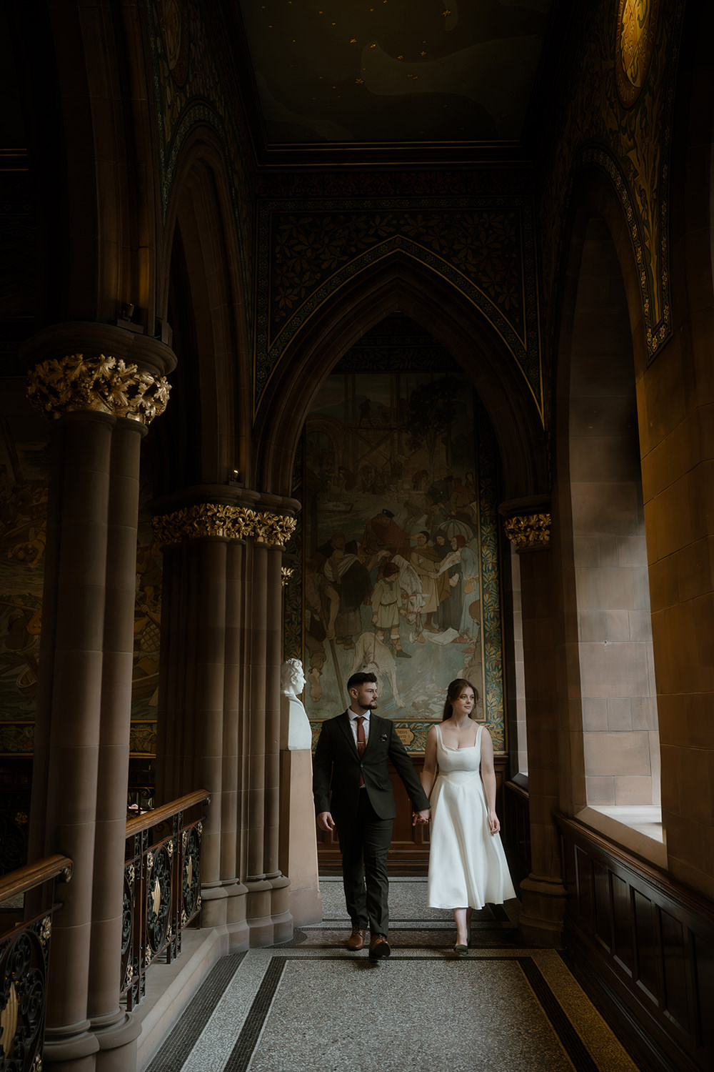 Newlyweds walk hand in hand through a Gothic-style corridor, surrounded by arched columns with gold detailing, decorative murals, and a bust sculpture, illuminated by natural light from tall windows