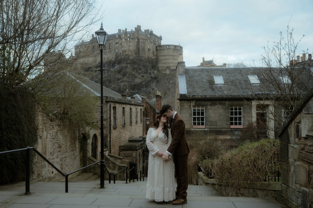 A couple stands close together on the Vennel steps in Edinburgh, with Edinburgh Castle prominently visible in the background. The historic stone buildings, slate roofs, and bare winter trees frame the scene