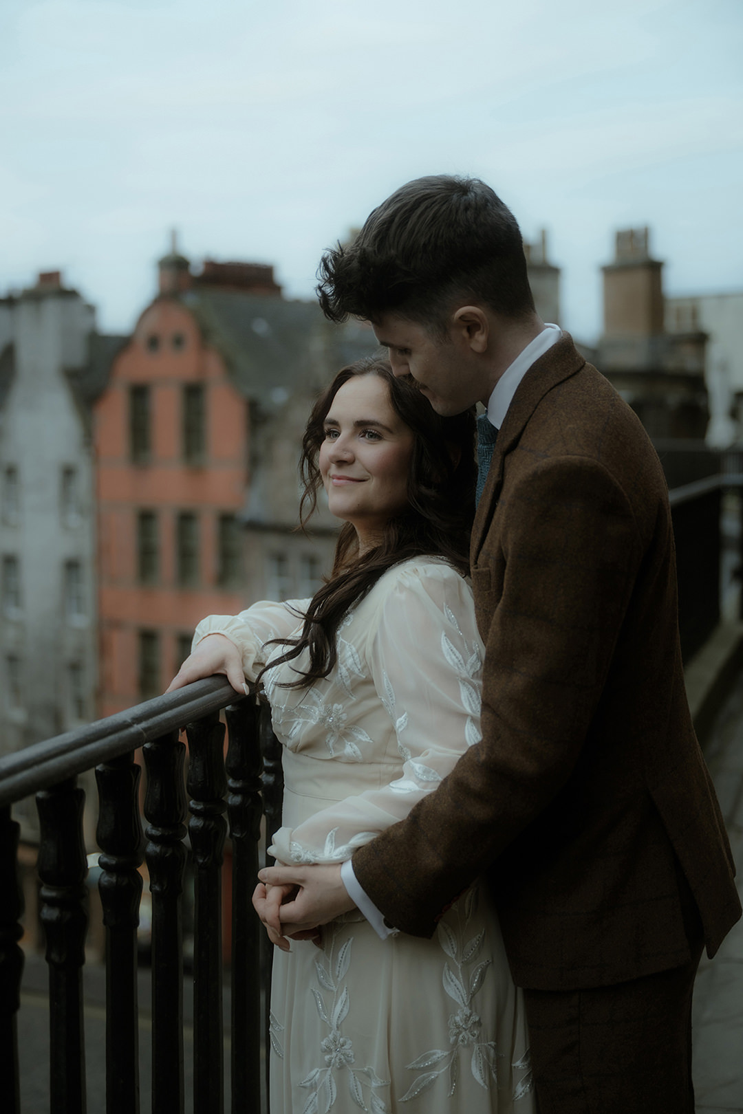 Bride and groom embrace on a wrought-iron balcony overlooking Old Town Edinburgh’s winter rooftops, the bride in a long-sleeve embroidered gown and the groom in a brown tweed suit