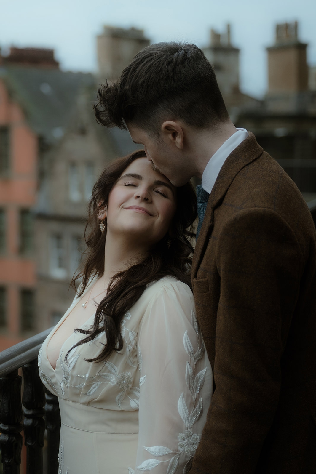Groom in a brown tweed suit kisses his bride’s forehead on a stone balcony in winter Edinburgh, her ivory leaf-patterned wedding dress and the historic city skyline visible behind them
