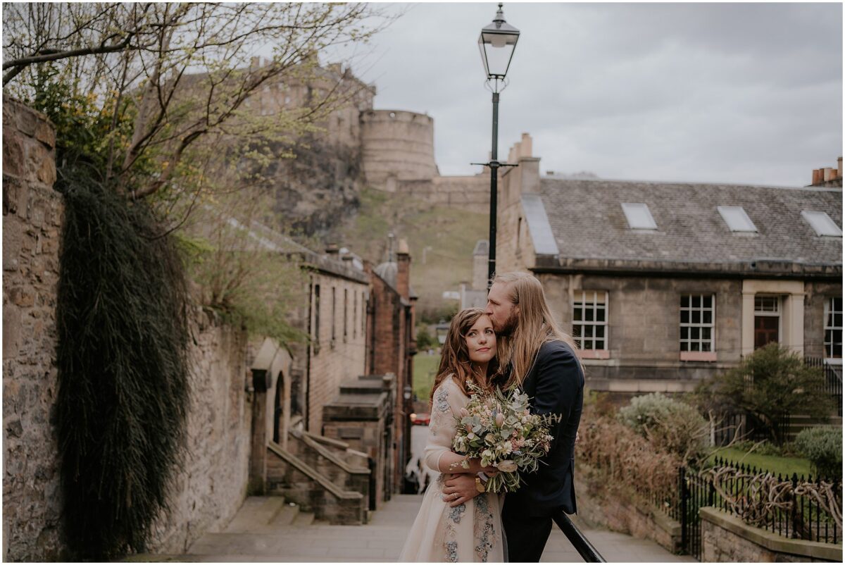 Groom kissing the bride on the forehead with Edinburgh Castle towering behind them