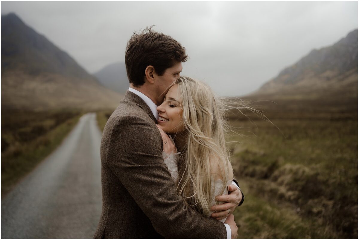 Tender embrace on Glen Etive road: bride nestles into groom's chest with eyes closed, cradled warmly in his arms as the empty road and mountains stretch beyond
