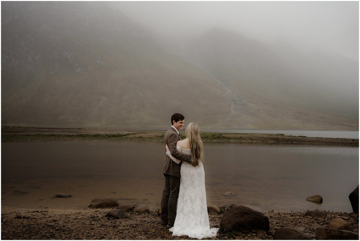 The bride in a flowing lace gown and the groom in a tweed jacket stand side by side at the edge of a still Loch Etive