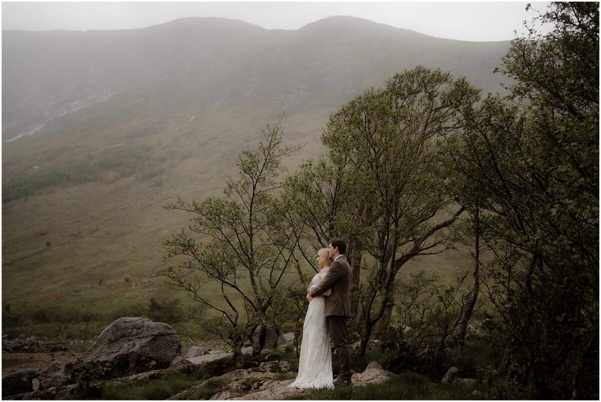 From behind, the groom and bride hold each other close on a pebbled shore
