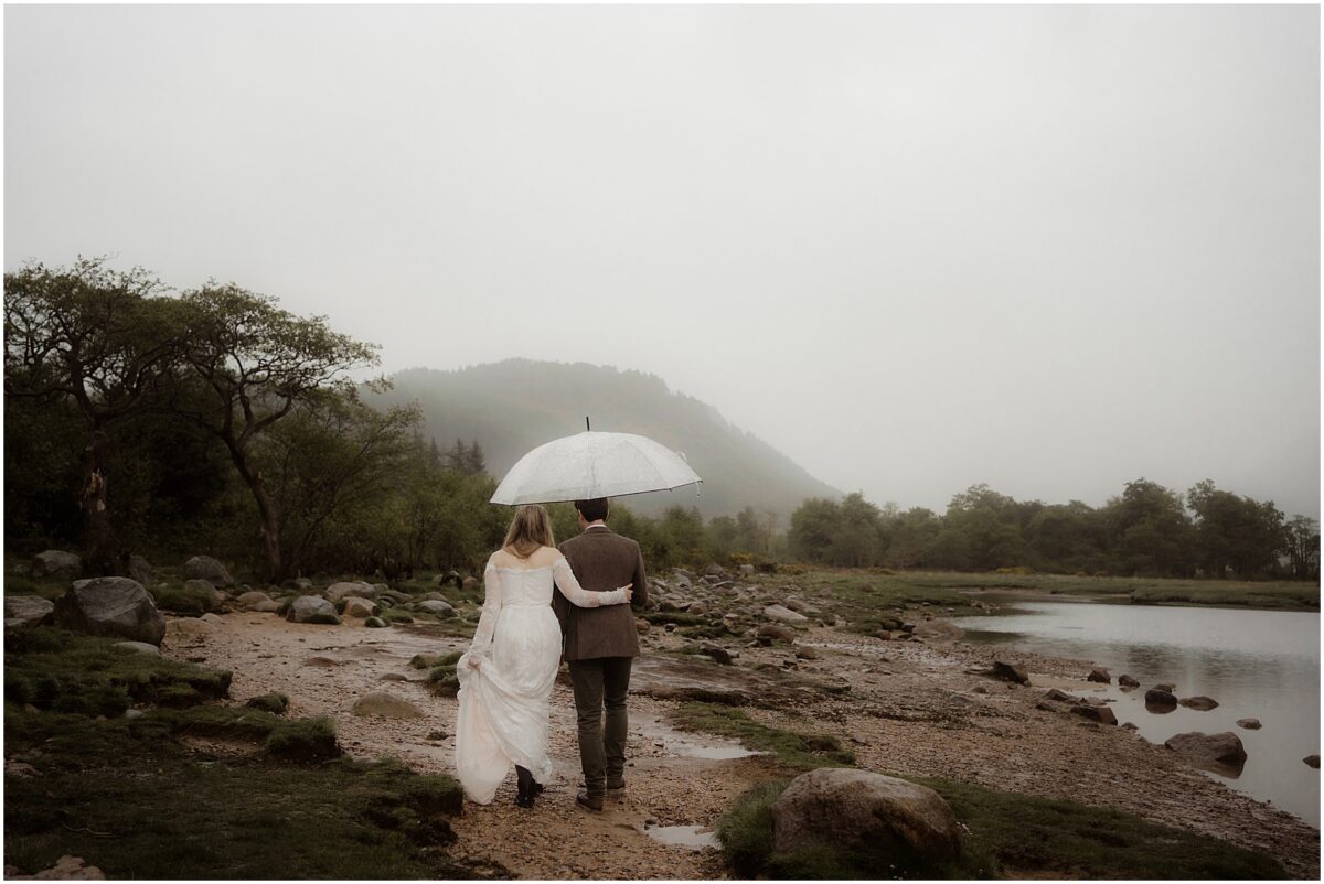 Bride and groom walk arm-in-arm along a pebbled riverside track beneath a clear umbrella