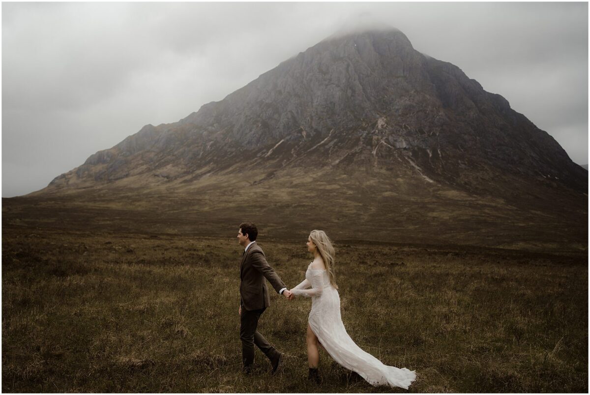 Groom leads bride by the hand across the windswept moor, her lace train trailing through the heather as Buachaille Etive Mòr’s craggy summit rises dramatically behind them