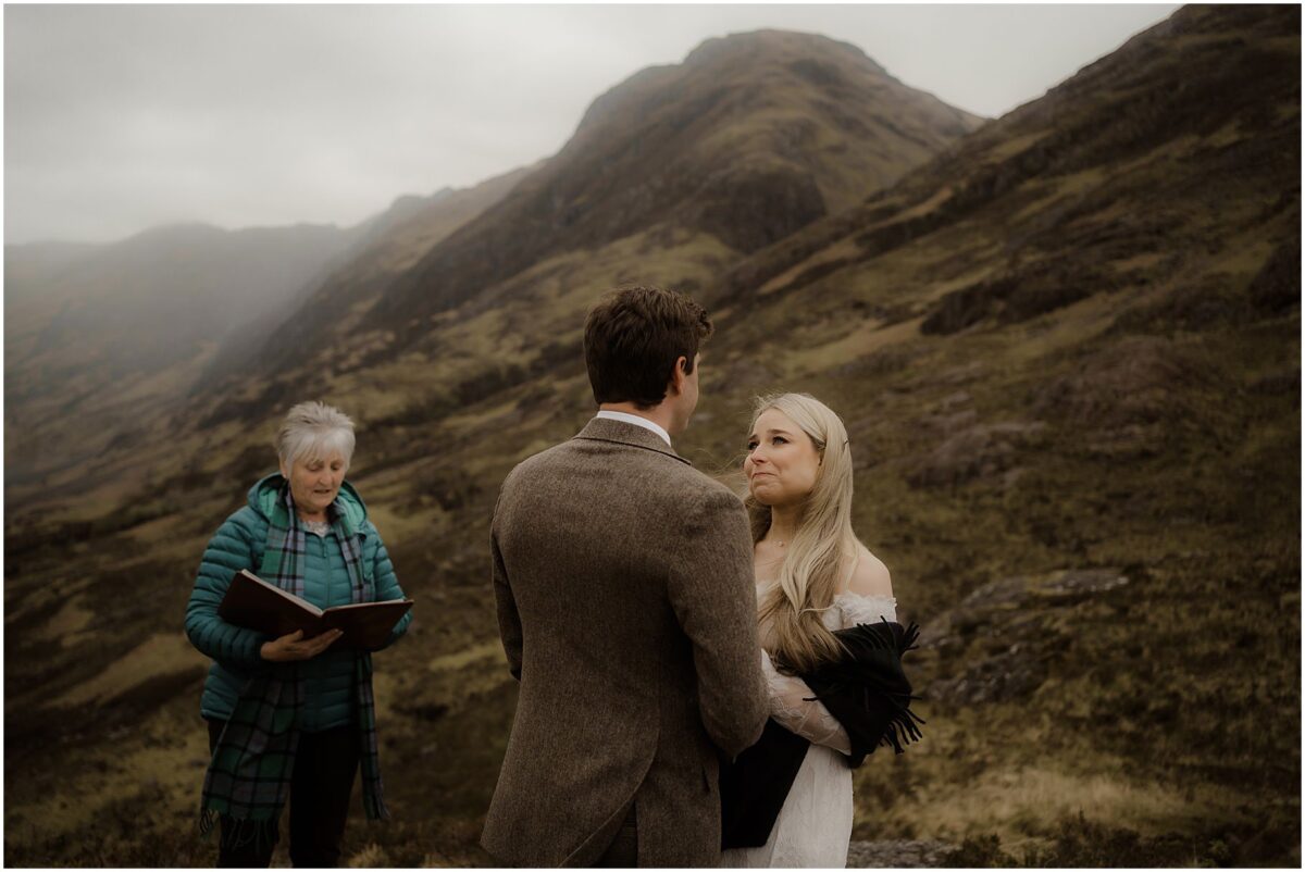 Bride and groom exchanging emotional vows atop a Glencoe peak, misty valley and winding road below
