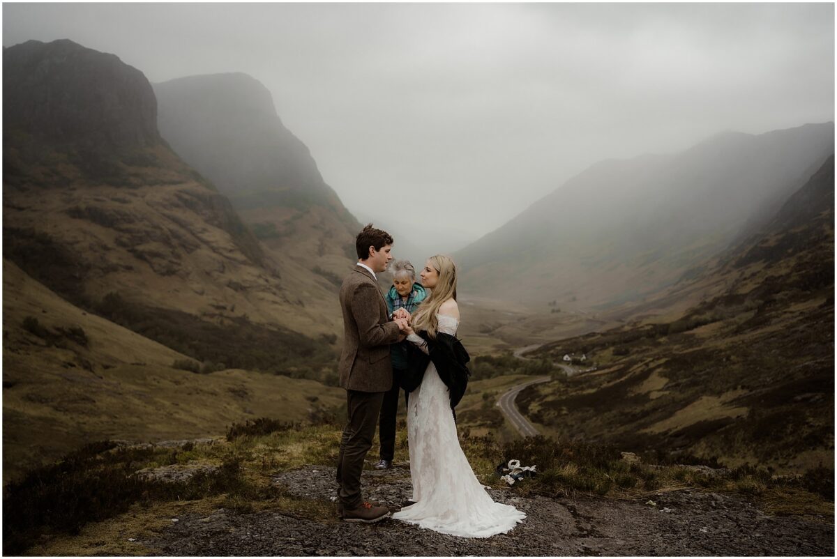 Wide shot of the handfasting ritual: officiant in tartan shawl binding the couple’s hands with Celtic cord, mountains looming behind