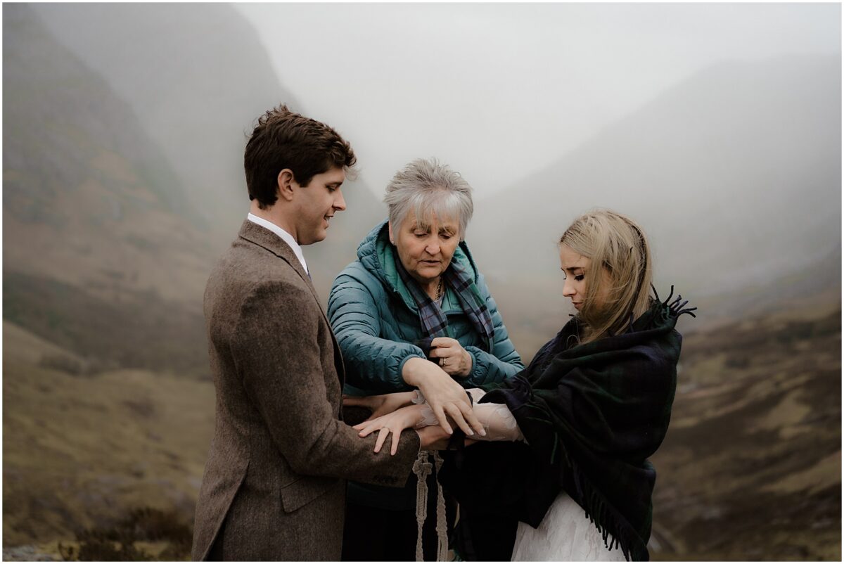 Bride and groom holding hands mid-ceremony, their officiant tying a braided cord around their wrists on a rocky outcrop