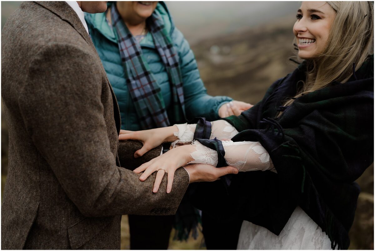 Officiant tightening the handfasting knot while the bride laughs, her veil and shawl fluttering in the wind.