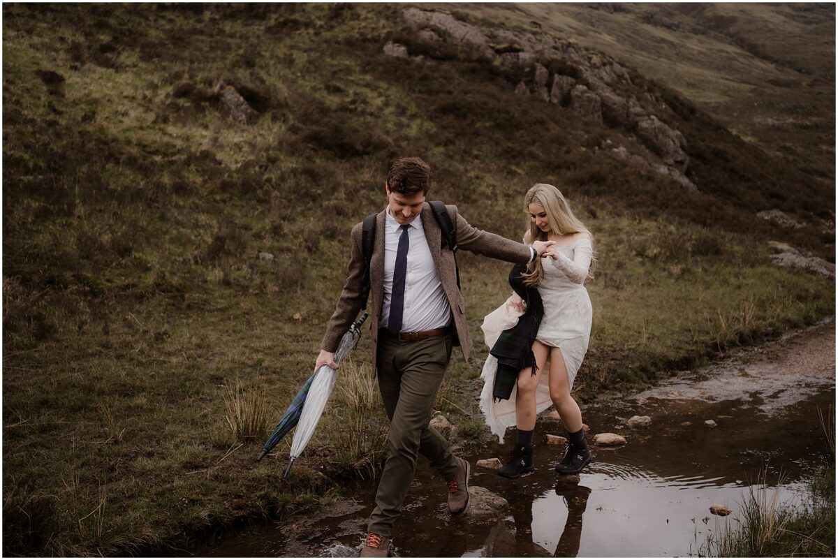 Groom leads bride across a shallow stream on a rocky Highland trail, both in hiking boots and wedding attire - hiking elopement in Scotland