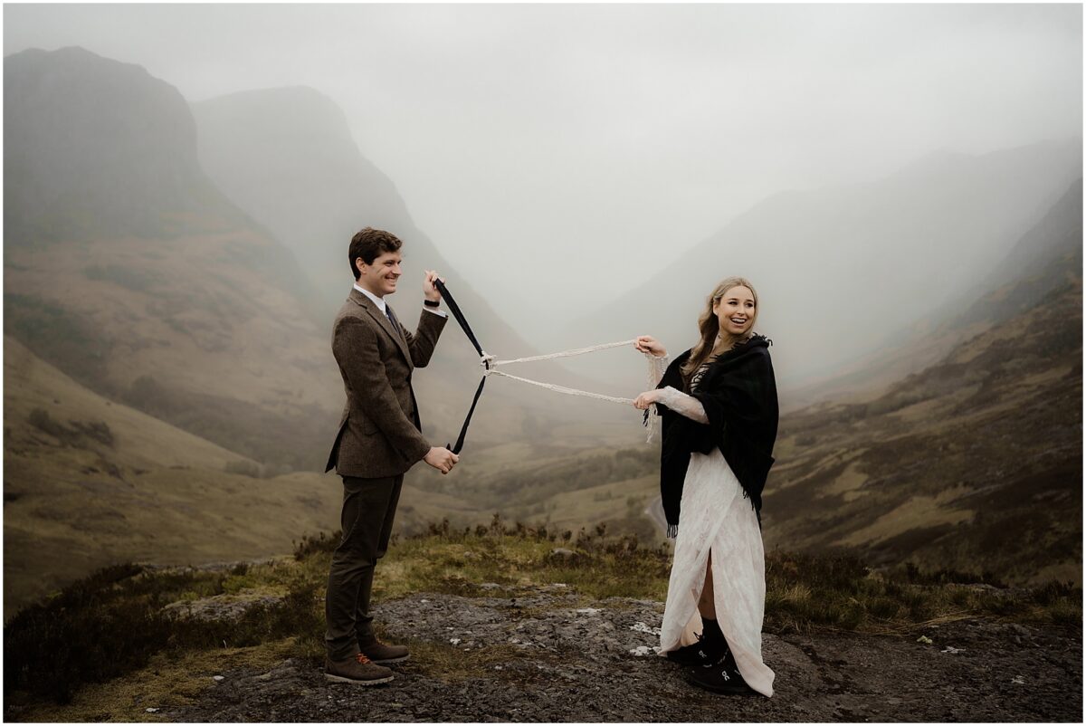 Couple tying the knot in the middle of their handfasting cords, with misty Scottish mountains surrounding them