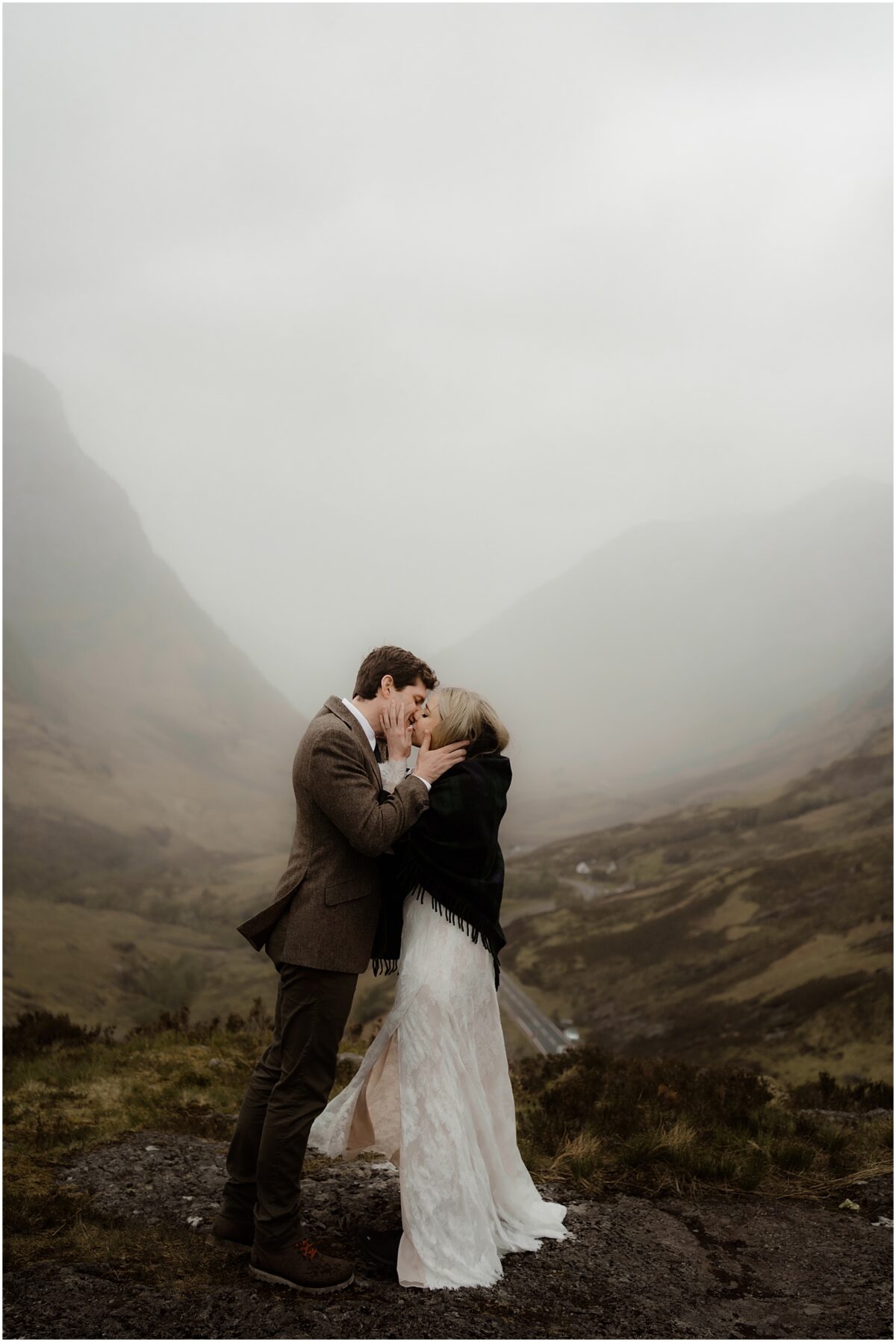 Newlyweds share a misty kiss on a rocky ledge, bride's white dress and shawl billowing in the breeze