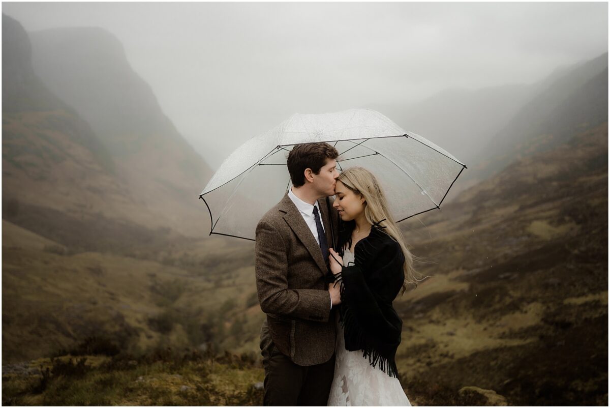 Groom kissing bride's forehead under a clear umbrella, raindrops glistening as the Glencoe valley looms behind