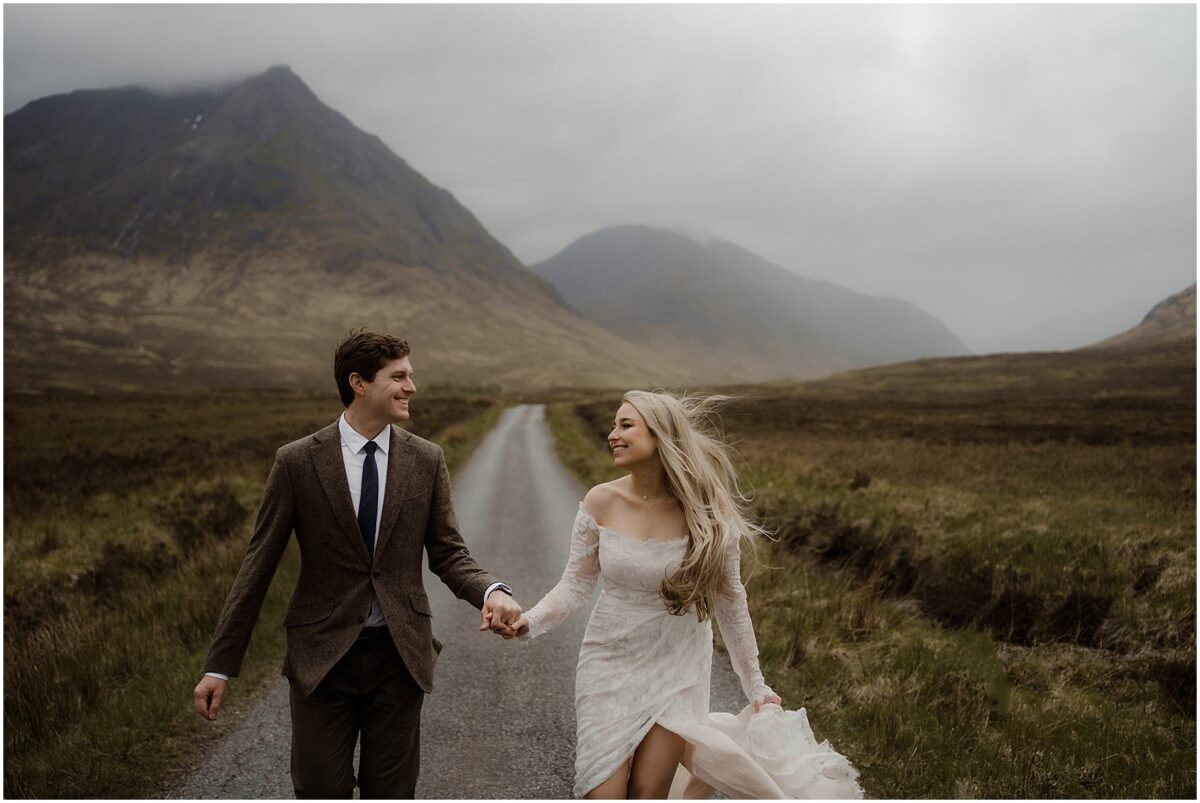 Newlyweds sprint hand-in-hand down a narrow Highland road, her lace dress billowing and their faces lit with laughter as rugged peaks rise behind them under brooding skies