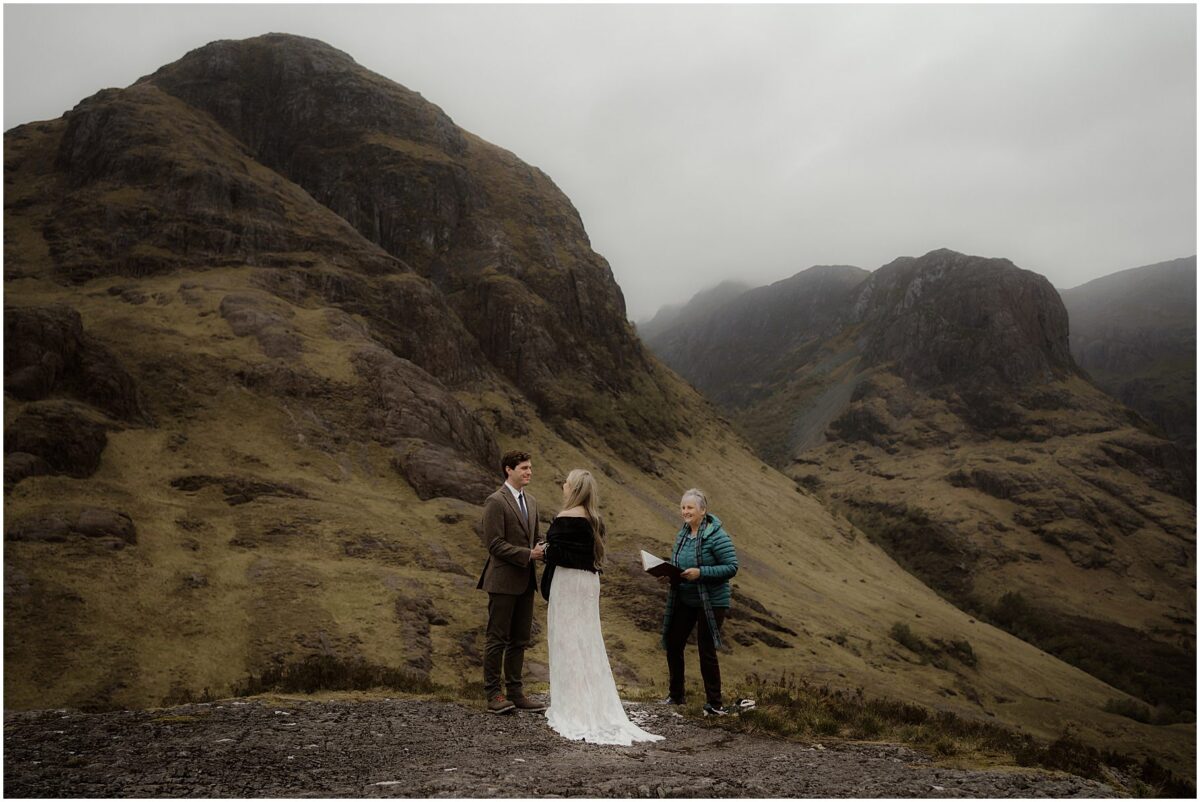 Wide-angle view of the couple and officiant at their mountain ceremony, with steep green ridges behind them