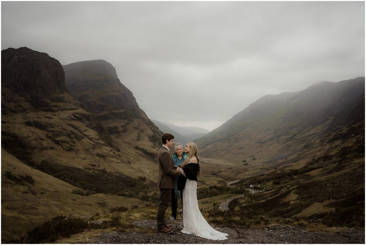 Soon-to-be-married couple stand hand in hand while their officiant reads vows on a windswept rock ledge in Glencoe