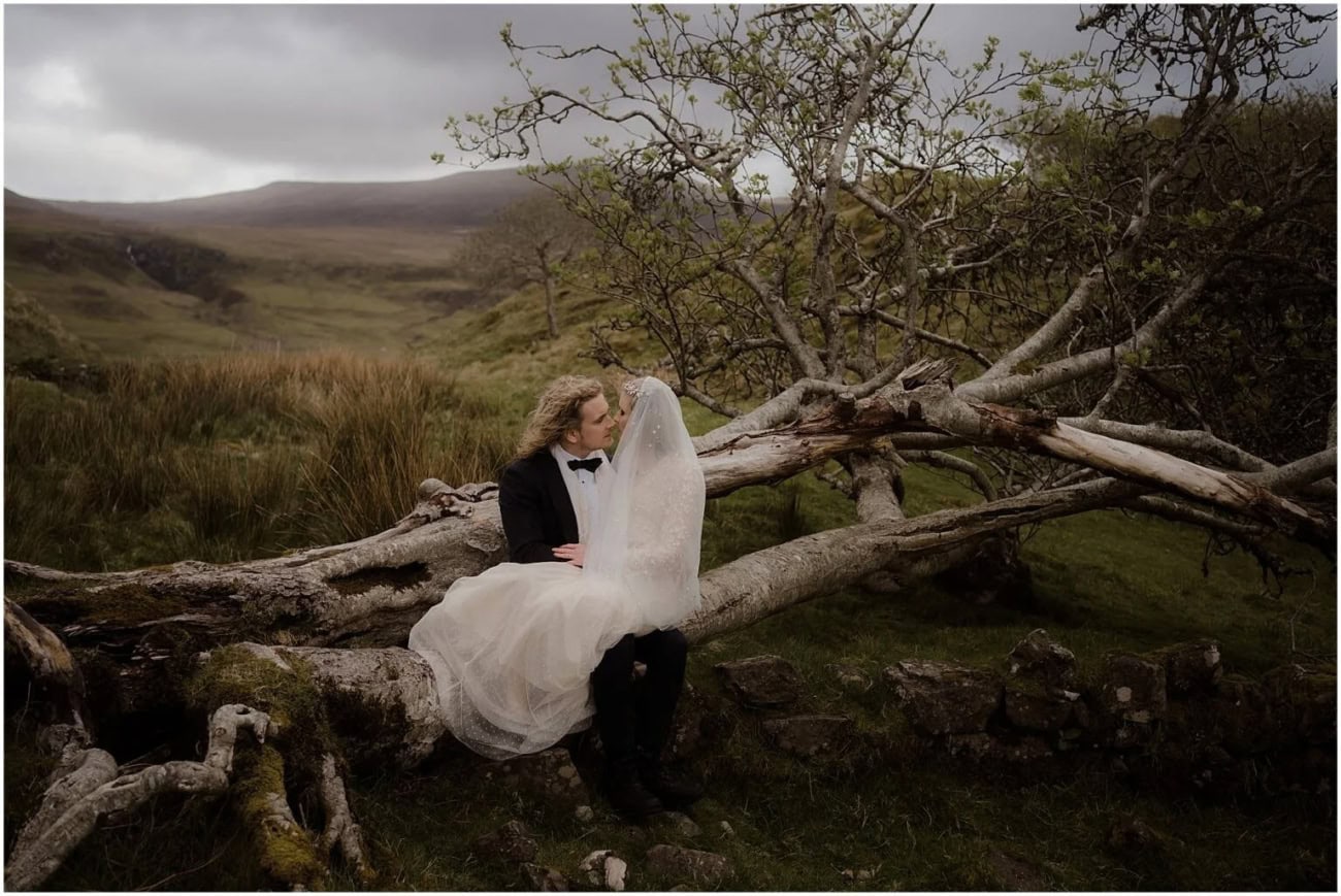 Rainy Fairy Glen elopement on Isle of Skye, Scotland ride and groom sitting together on a fallen tree trunk amid the rugged green highlands of the Isle of Skye, the bride's veil caught in the wind