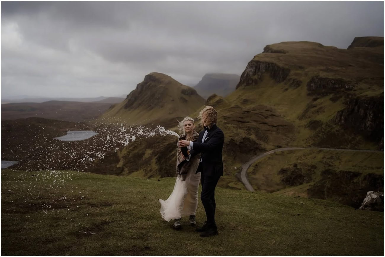 Rainy Fairy Glen elopement on Isle of Skye, Scotland Groom drinking champagne from the bottle while bride laughs beside him on the Quiraing ridge
