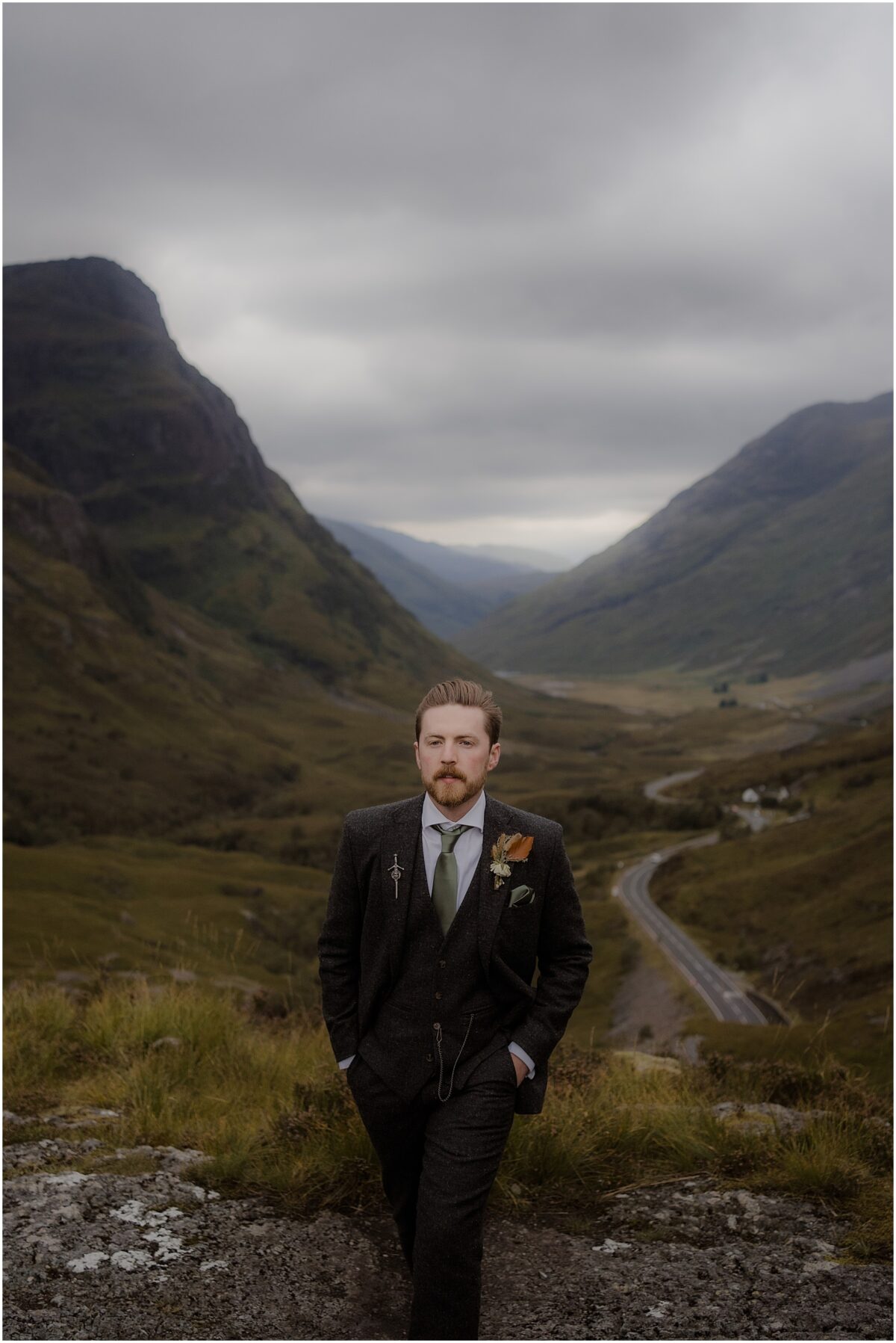 Wedding photos in Glencoe - Glencoe wedding photographer Portrait of the groom wearing tweed suit and a light green tie, hands in his pockets and a winding highland road behind him
