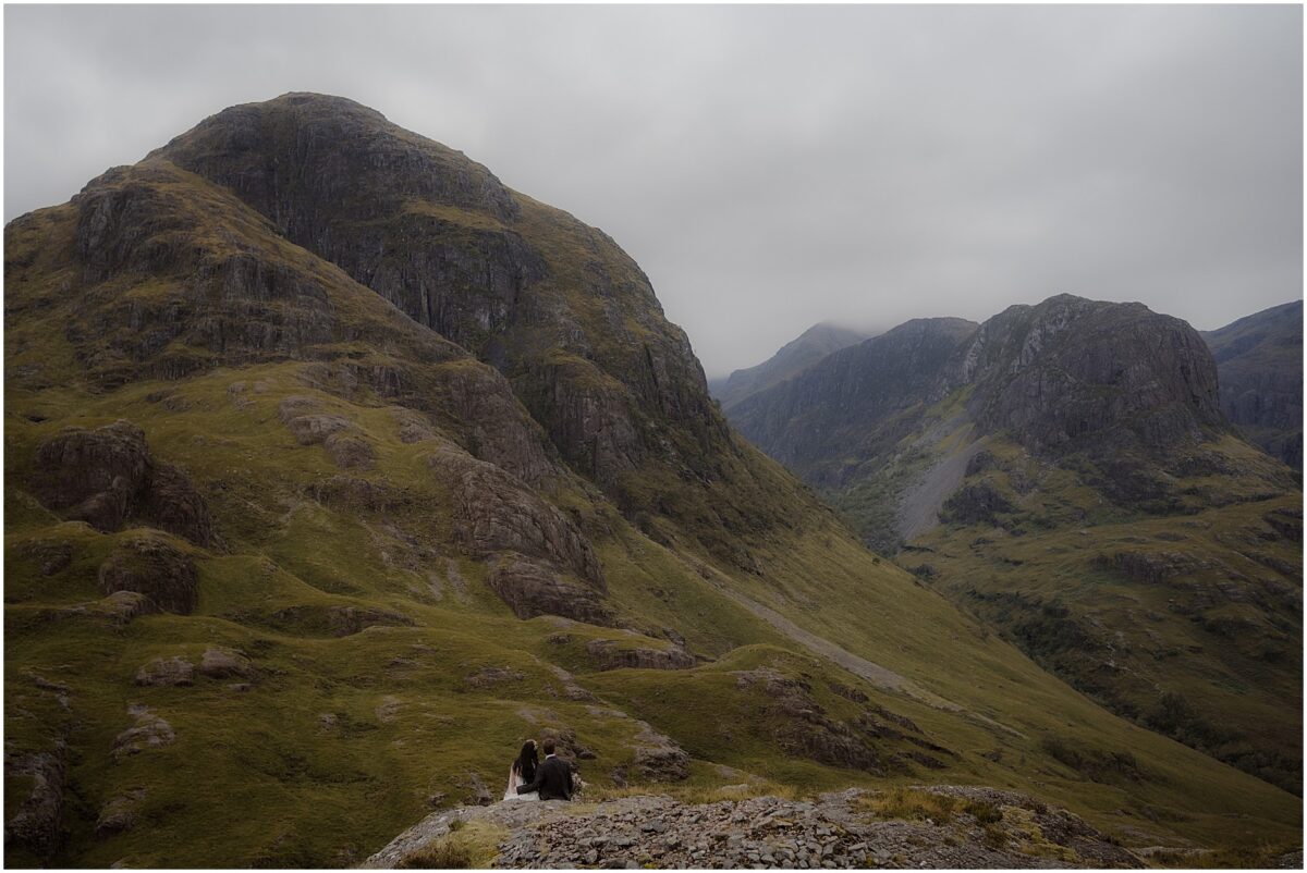 Wedding photos in Glencoe - Glencoe wedding photographer Wide-angle view at Glencoe Valley, little silhouettes of bride and groom sitting side by side at the bottom of the mountain