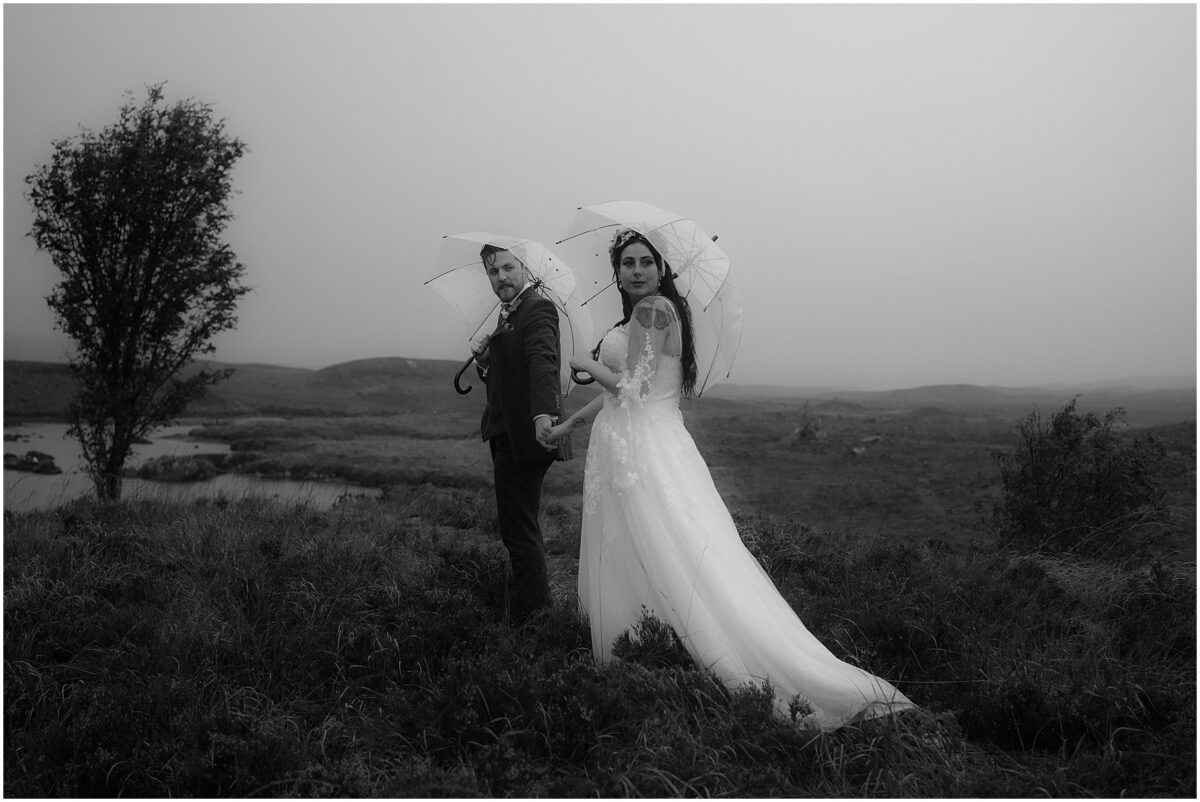 Wedding photoshoot in Glencoe - Glencoe wedding photographer Couple walking towards Rannoch Moor holding umbrellas and looking over their shoulders, in black and white