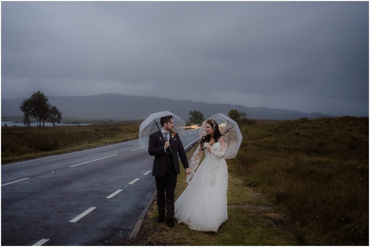 Wedding photoshoot in Glencoe - Glencoe wedding photographer Bride and groom walk by the road holding umbrellas in the rain, with backlit car headlights behind them