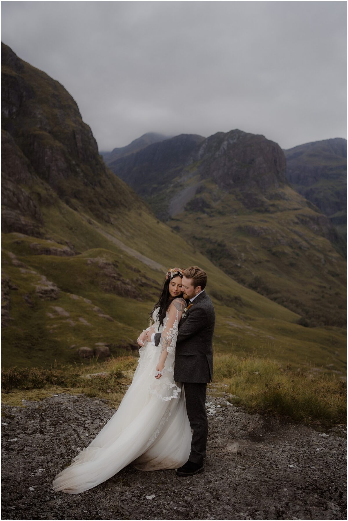 Glencoe wedding photos - Glencoe wedding photographer Close embrace of the newlyweds - her in a long wedding dress and a flower crown and him in a dark tweed suit, with Three Sisters of Glencoe behind them