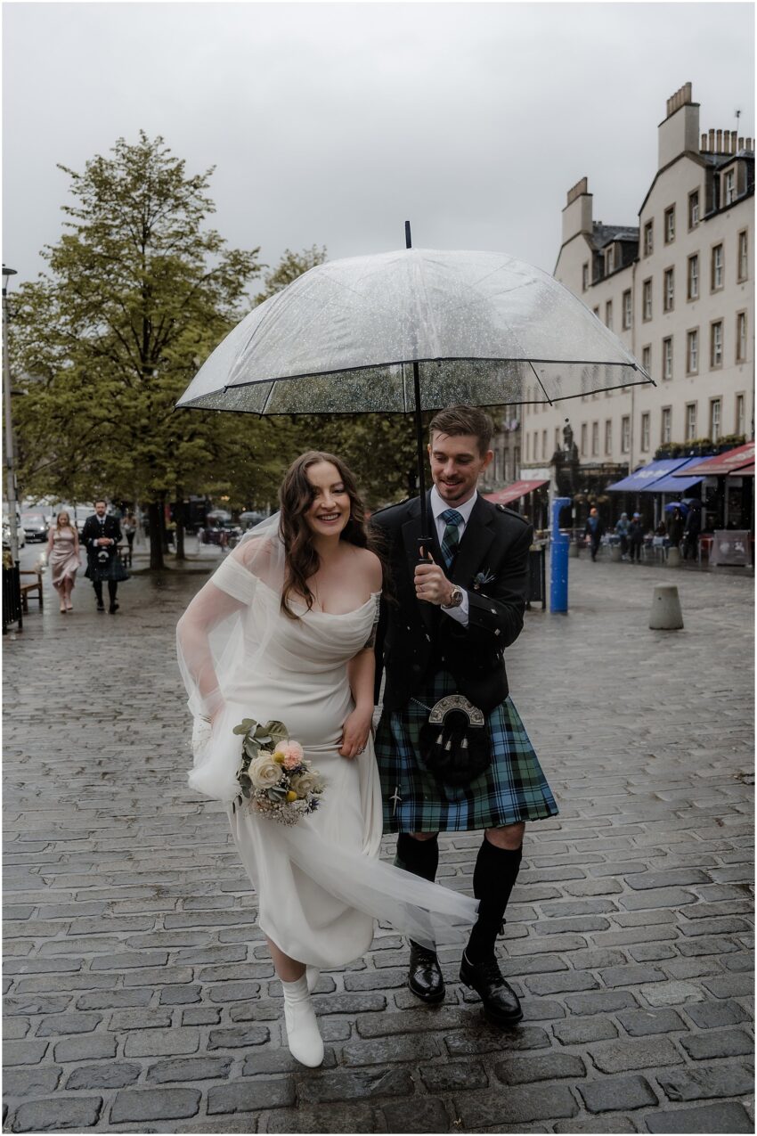 Bride and groom walking underneath a clear umbrella on the Grassmarket, Edinburgh