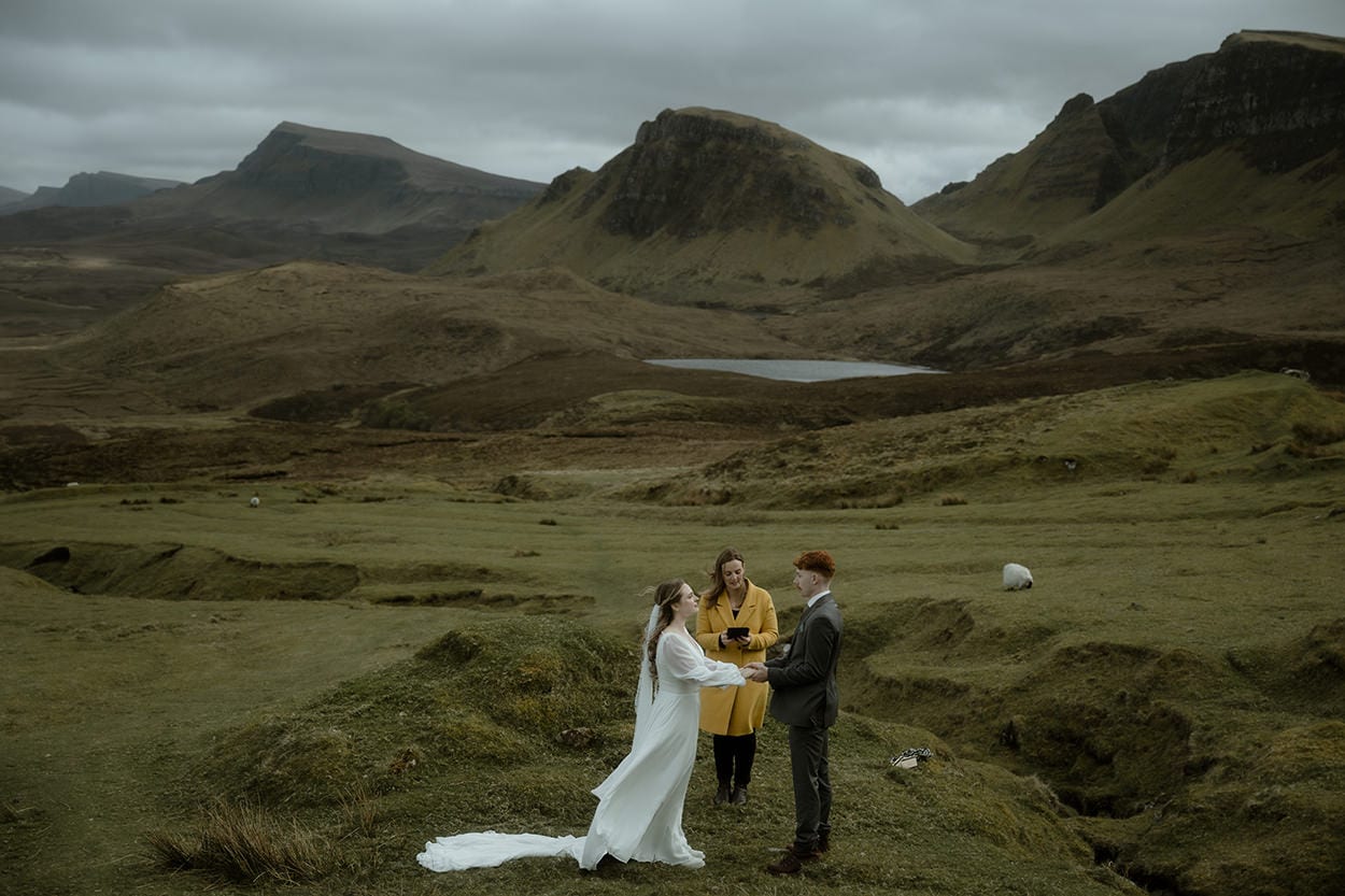 Bride and groom holding hands during their elopement ceremony at the Quiraing, Isle of Skye, with a celebrant officiating - a loch, rolling green hills, and grazing sheep stretching into the distance behind them