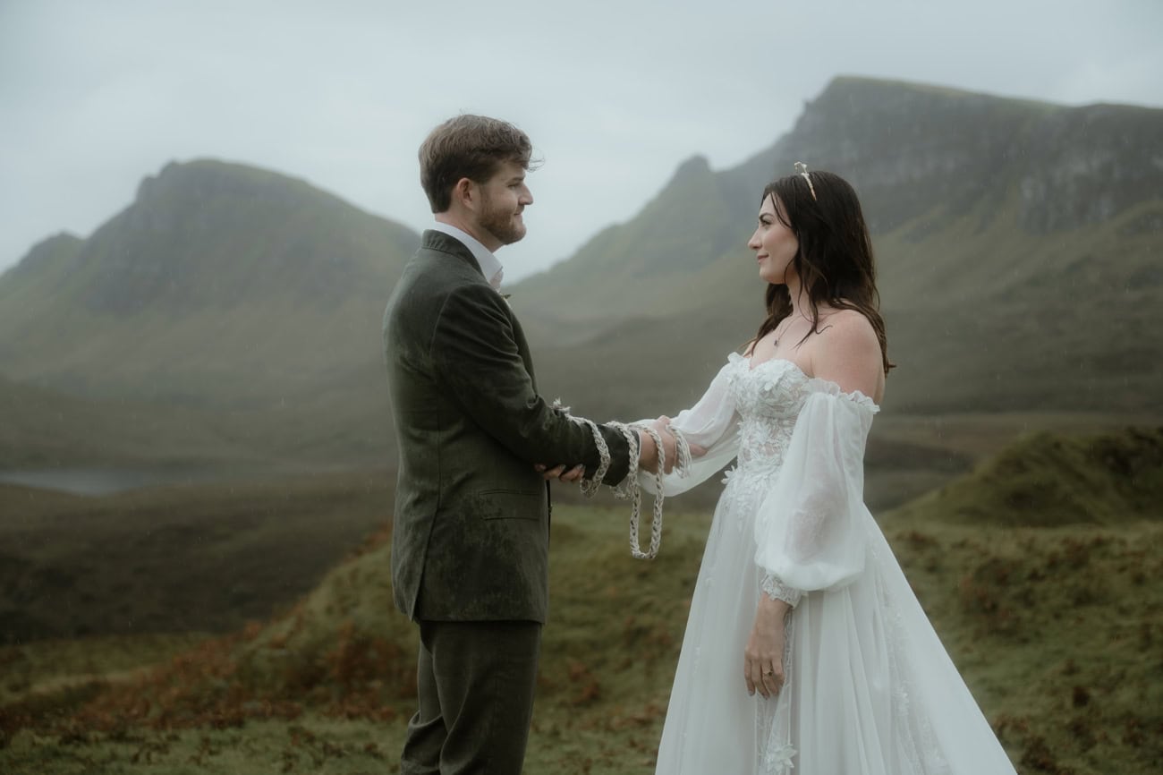 Isle of Skye Elopement Guide 2026/27 – plan your Scottish adventure wedding 1 Eloping couple during a handfasting ceremony at the Quiraing, Isle of Skye, hands bound together with cord, dramatic misty peaks rising behind them