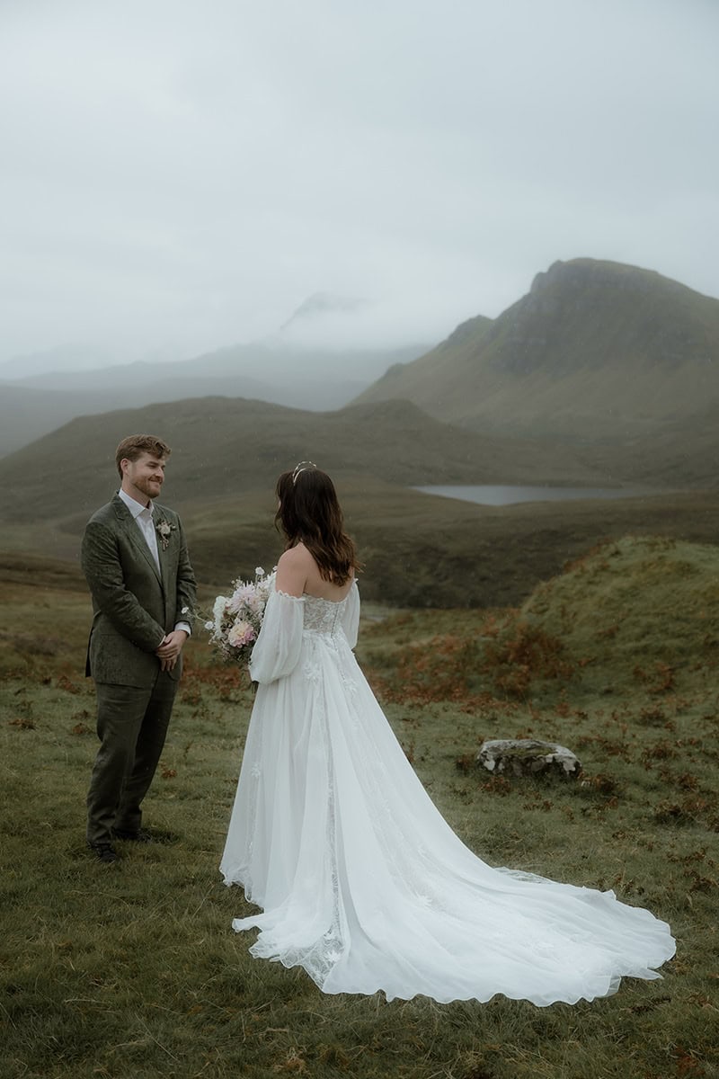 Isle of Skye Elopement Guide 2026/27 – plan your Scottish adventure wedding 2 Bride in an off-shoulder lace gown with a trailing dress facing her groom on the misty hillside at the Quiraing, Isle of Skye, with a loch visible in the valley below