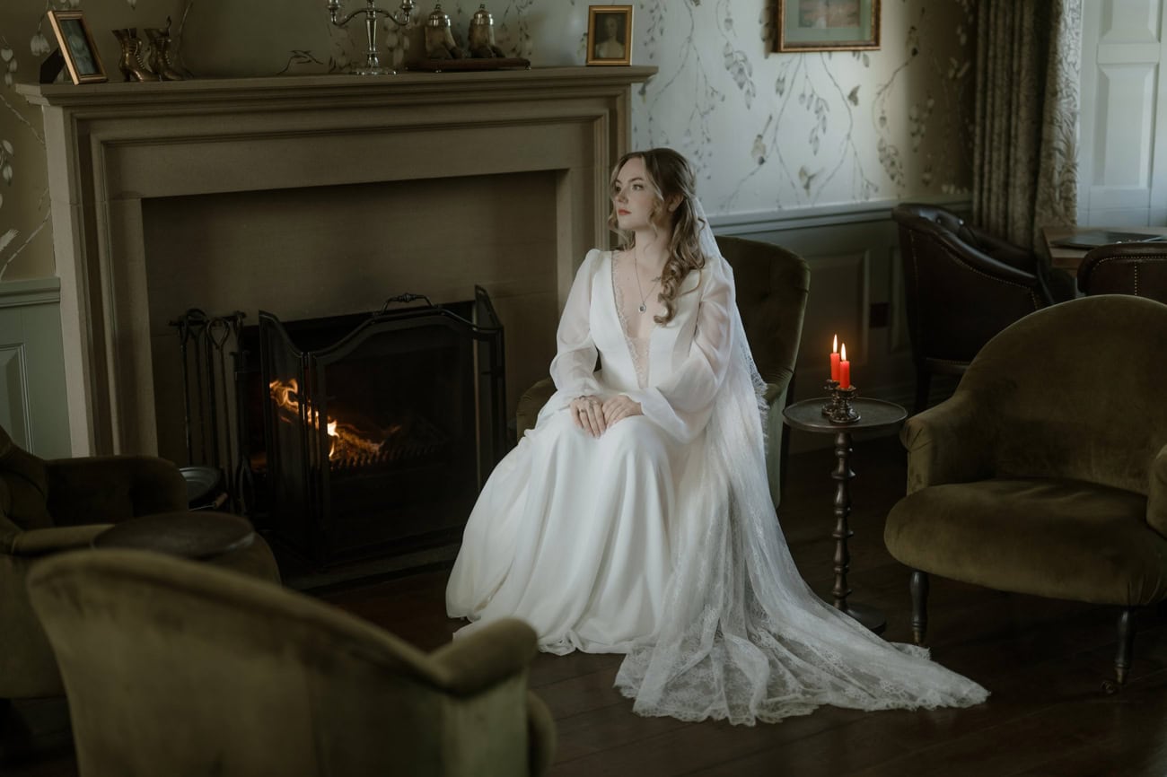 Isle of Skye Elopement Guide 2026/27 – plan your Scottish adventure wedding 28 Bride in a deep V-neck silk wedding gown sitting beside a lit fireplace at Monkstadt 1745 hotel, Isle of Skye, lace veil trailing across the floor, red candle burning nearby