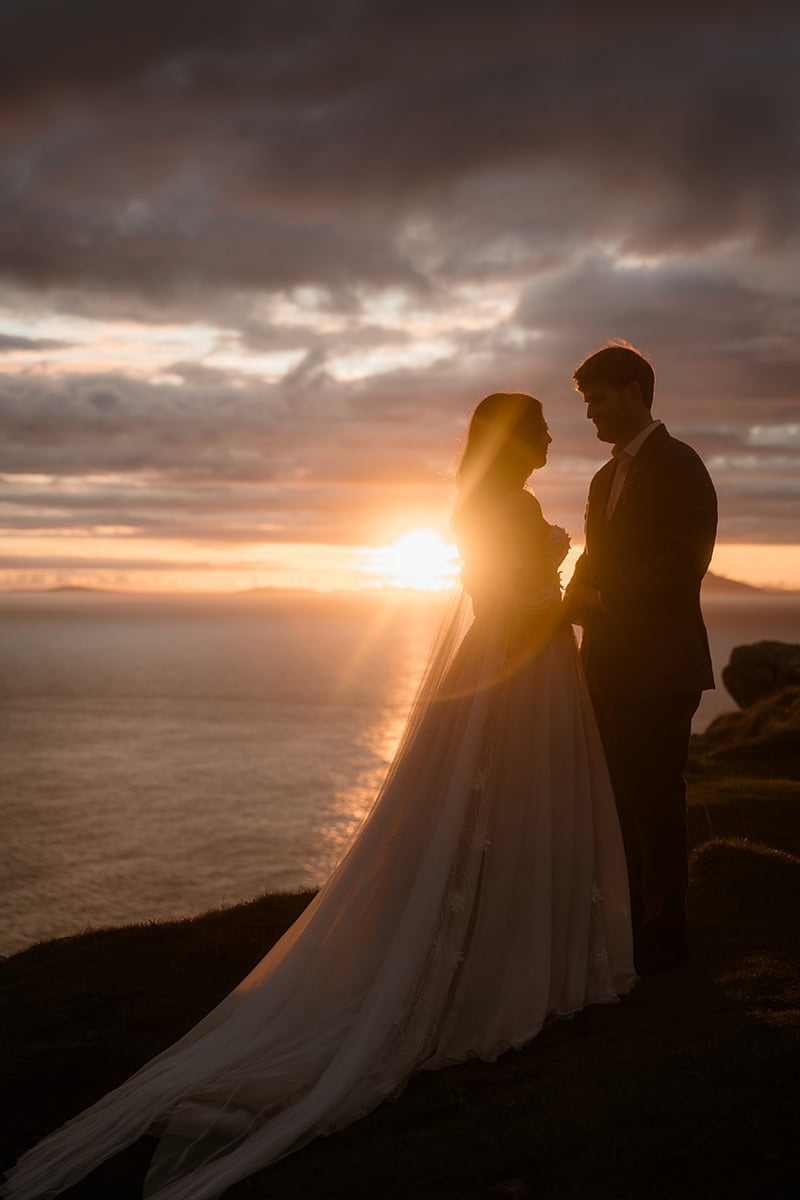 Isle of Skye Elopement Guide 2026/27 – plan your Scottish adventure wedding 14 Silhouette of a couple facing each other at Neist Point, Isle of Skye, with the sun setting directly behind them over the Atlantic, bride's veil and dress trail visible against the warm sky