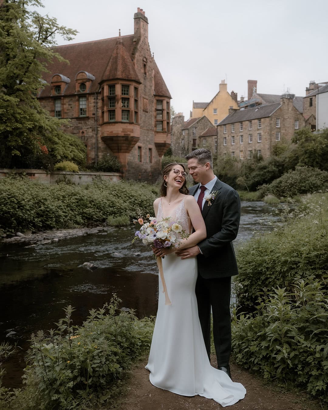 Micro-Wedding at Edinburgh Royal Botanic Garden 58 Bride in a slip gown laughing with groom in a dark suit beside the Water of Leith in Dean Village, lush summer greenery and the iconic Well Court building reflected in the river behind them — Royal Botanic Garden Edinburgh micro-wedding photography by Natalia Swiader