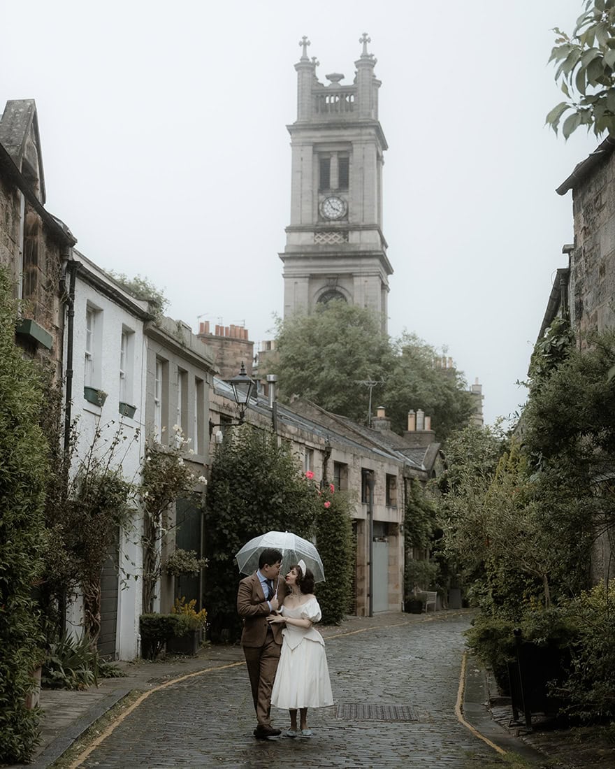 Intimate dark academia inspired Edinburgh wedding 64 Bride and groom share a kiss under a clear umbrella on a rainy day in Circus Lane, Edinburgh, surrounded by rose-covered stone cottages and St Stephen's Church tower rising through the mist