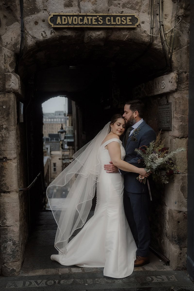 Bride in a fitted off-shoulder gown with a flowing veil and groom in a navy suit sharing an intimate embrace beneath the stone archway of Advocate's Close on the Royal Mile, Edinburgh