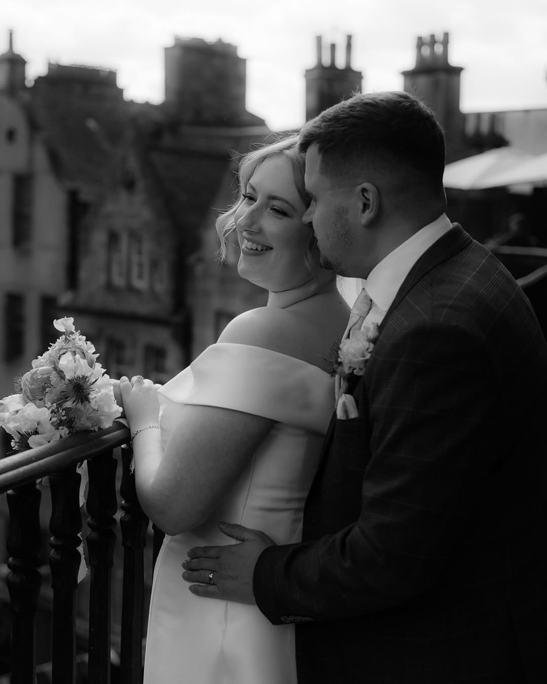 Bride in an off-shoulder gown and groom in a checked suit sharing an intimate moment on Victoria Terrace, Edinburgh Old Town rooftops softly blurred behind them