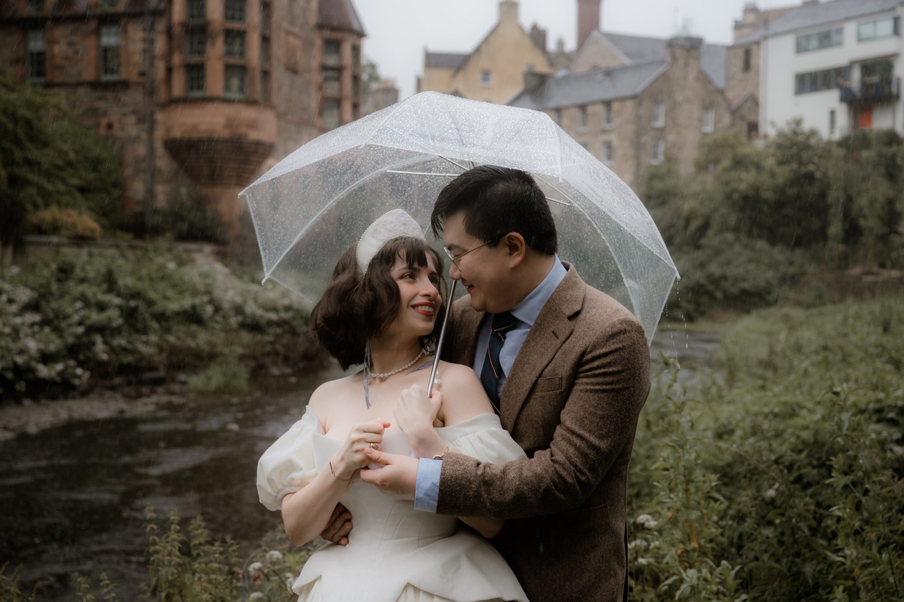 Advice for Rainy Wedding Days in Scotland - turn rain into romance 4 Bride and groom underneath an umbrella, smiling at each other with Edinburgh Dean Village in the background