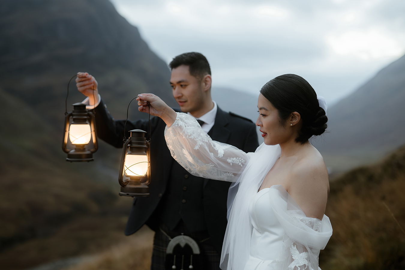 A newlywed couple stands against the dramatic backdrop of Glencoe, Scotland, each holding a glowing lantern. The bride, dressed in an ethereal off-the-shoulder gown with delicate lace sleeves, smiles as she lifts her lantern. The groom, wearing traditional Scottish attire, gazes at her warmly. The soft light from the lanterns contrasts with the moody, misty mountains behind them