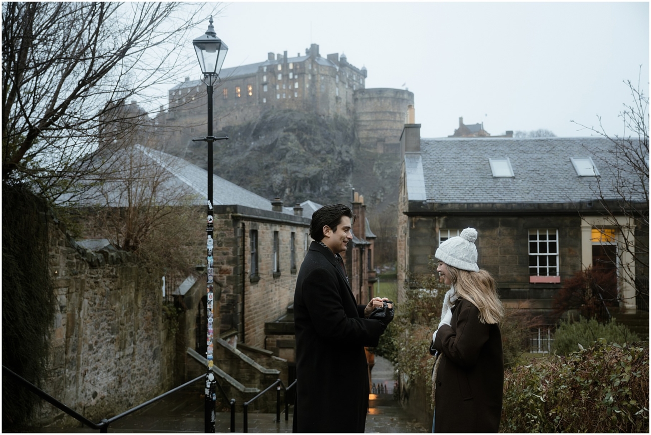 The man holds an engagement ring box while the woman smiles warmly. Edinburgh Castle towers dramatically in the misty background, adding a magical atmosphere to the proposal.