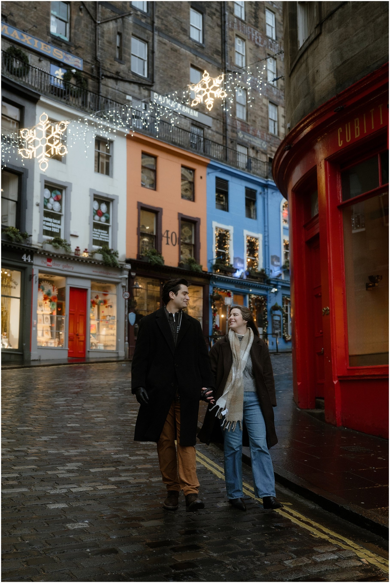 A vibrant engagement photo on Victoria Street, featuring the couple walking hand-in-hand, surrounded by brightly coloured shopfronts and festive holiday lights strung across the street