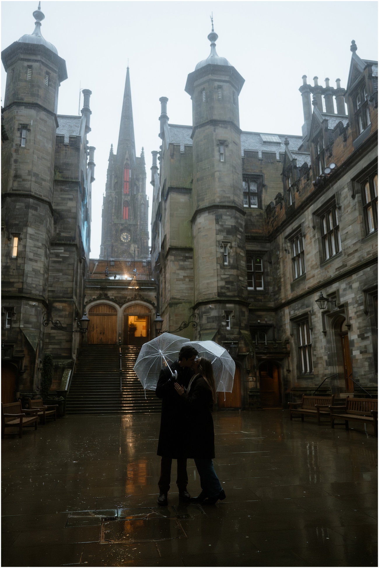 A romantic shot of a couple embracing under a clear umbrella in the courtyard of Edinburgh's historic Old Town at the Divinity College, with a stunning gothic tower in the background and rain glistening on the stone pavement