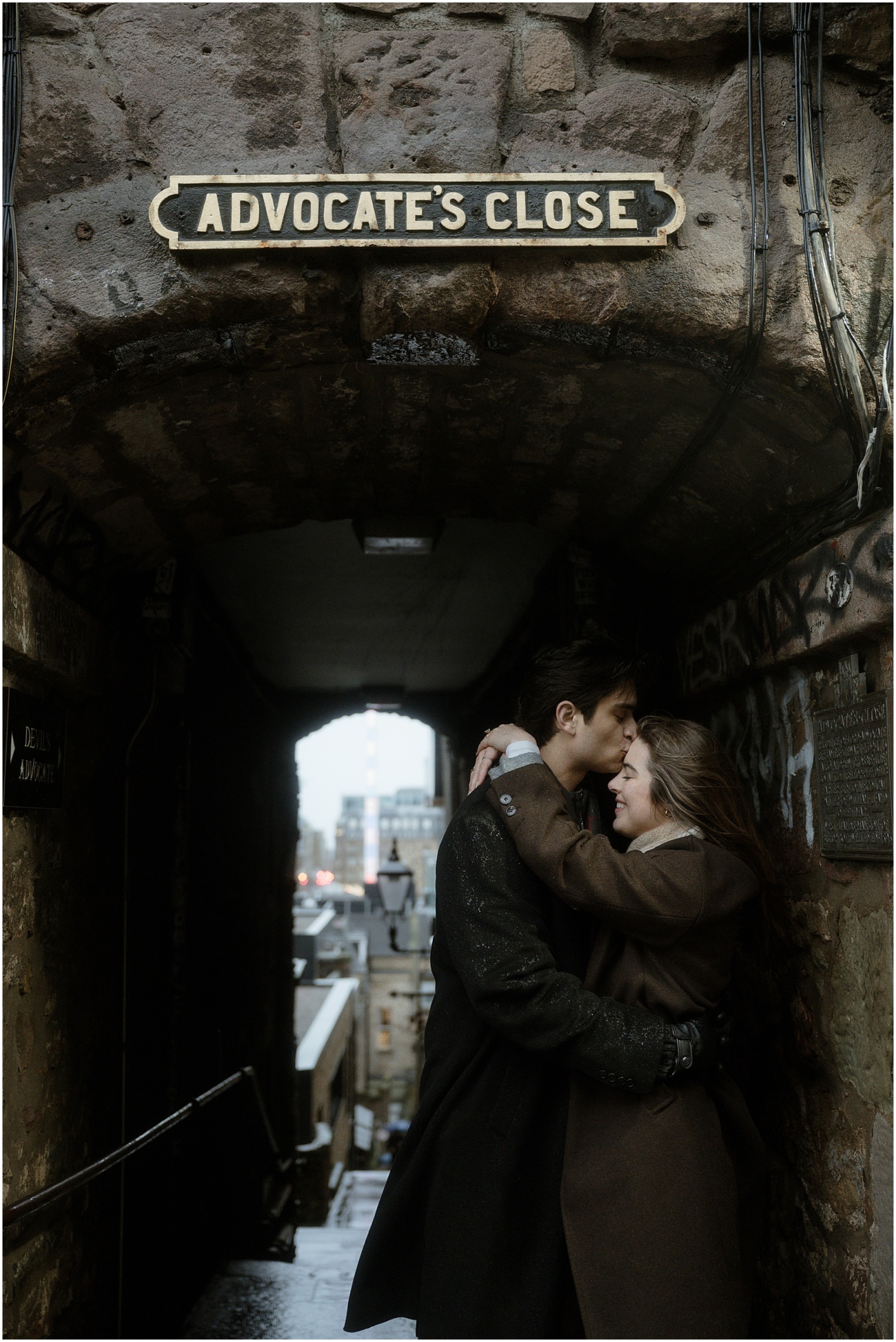 The couple standing beneath the "Advocate's Close" sign in Edinburgh, sharing an intimate moment with the bustling cityscape blurred in the background