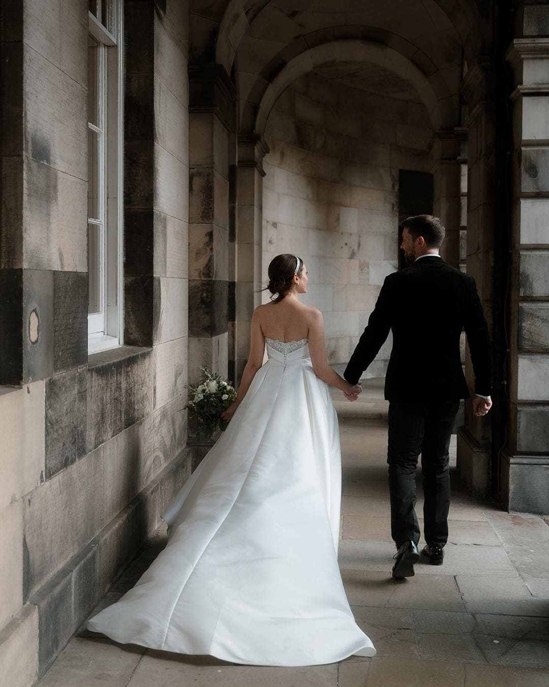 Elegant black tie Bonham Hotel Wedding in Edinburgh 46 Bride in a strapless ballgown with a sweeping train and groom in black tie walking hand in hand through the stone archways of Edinburgh City Chambers