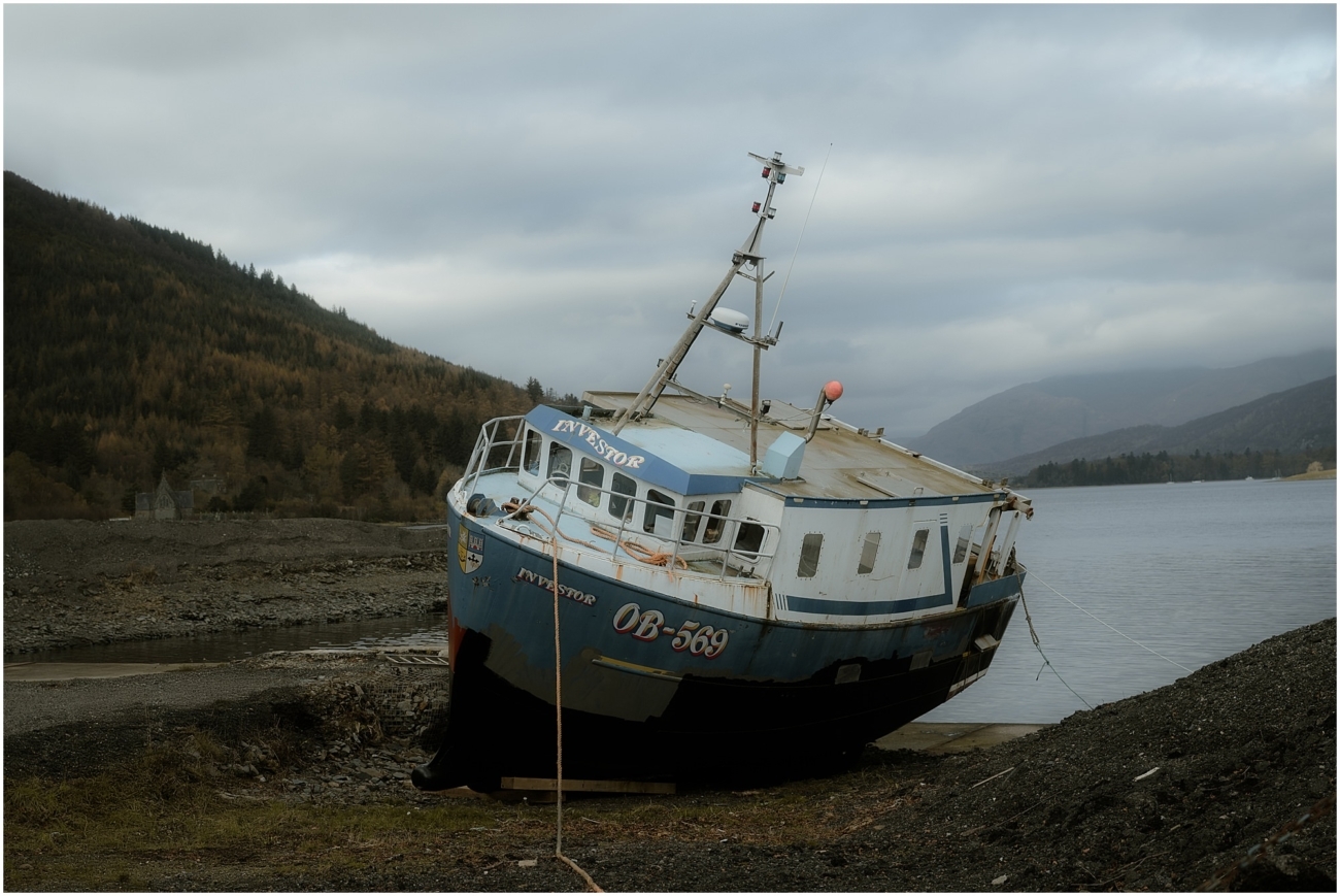 A beached blue fishing boat named 'Investor' resting on the rocky shore, with a scenic Highland loch and misty mountains in the background.